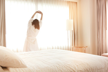 Woman stretching in bed after waking up, back view. Woman sitting near the big white window while stretching on bed after waking up with sunrise at morning, back view.