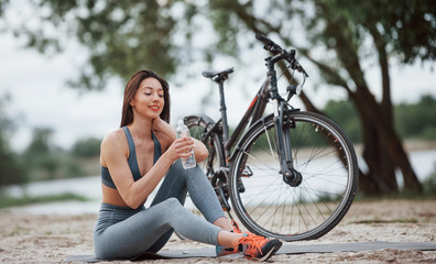 Obraz na płótnie Canvas Drinking water. Female cyclist with good body shape sitting near her bike on beach at daytime