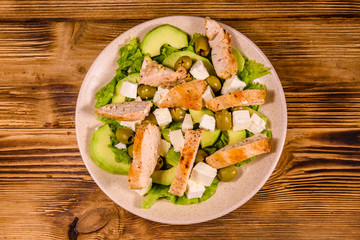 Fresh salad with chicken meat, feta cheese, avocado, green olives and lettuce leaves in ceramic plate on wooden table. Top view