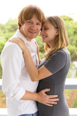 Fototapeta premium Happy expectant parents young posing on terrace. Man and pregnant woman standing outside, hugging each other, talking, looking at camera. Happy family couple concept