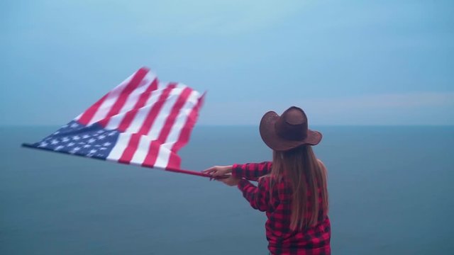Girl waving the American flag from side to side. She is wearing a cowboy hat and a red shirt on the background of the sea. The concept of patriotism.