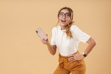 Image of amazed woman wearing eyeglasses smiling and holding smartphone