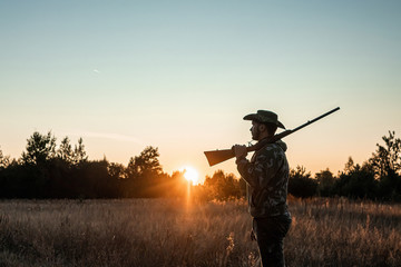 Silhouette of a hunter in a cowboy hat with a gun in his hands on a background of a beautiful sunset. The hunting period, the fall season is open, the search for prey.