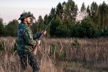 Fototapeta premium Hunter man in camouflage with a gun during the hunt in search of wild birds or game on the background of the autumn forest. Autumn hunting season. The concept of a hobby, killing.