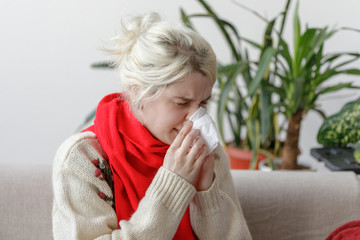Girl in a sweater sneezes while sitting on a sofa. The patient caught a cold, feeling sick and sneezing in a paper napkin.