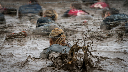 A woman crawling under barbed wire at a mud run  - obrazy, fototapety, plakaty