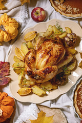 Thanksgiving or Halloween dinner with baked chicken with potatoes and lemons on big ceramic dish, pumpkin pies, plates, yellow autumn leaves, pumpkins as decorations over white table cloth. Flat lay