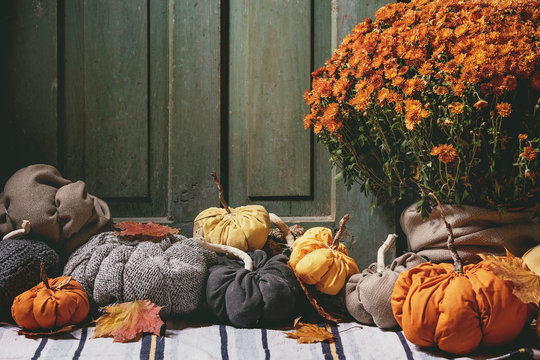 Wool and textile pumpkins for Halloween or Thanksgiving holidays home decor with autumn leaves, pot of chrysanthemums flowers on knitted doormat with old wooden door at background.