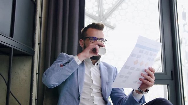 Agreeable clever bearded office manager drinking coffee from paper cup while reading financial report in the separate area in office