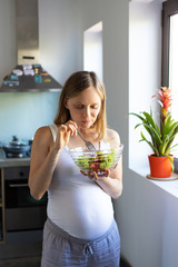 Excited expectant mother keeping healthy diet. Pregnant woman standing in kitchen, holding bowl, eating salad. Organic food concept