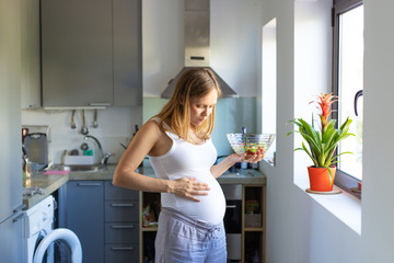 Excited expectant mother touching belly while enjoying organic lunch. Pregnant woman standing in kitchen, holding bowl, eating salad. Prenatal care and nutrition concept