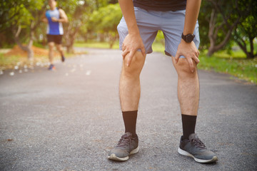 Male runner athlete knee injury and pain. Man suffering from painful knee while running in the public park.