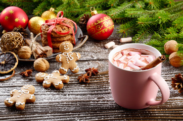 Christmas background of pink mug of hot chocolate with marshmallows, spruce branch and tray with gingerbread cookies on wooden table