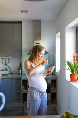 Positive expectant mother excited with organic lunch. Pregnant woman standing in kitchen, holding bowl, eating salad. Prenatal care and organic nutrition concept