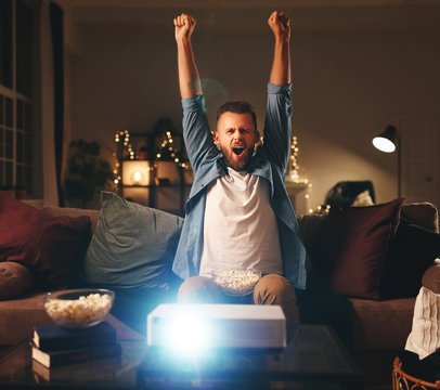 young male cheerleader watching football on tv projector at home in evening.