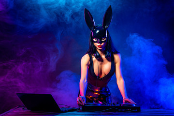 Young sexy woman dj playing music in mask. Headphones and dj mixer on table. Smoke on background