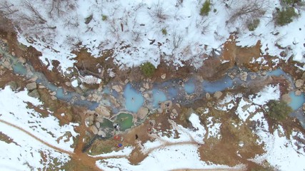 AERIAL TOP DOWN: Unrecognizable tourists enjoy a relaxing bath in the hot spring pools in snowy Colorado. Flying along a turquoise colored stream crossing the wintry landscape serving as a natural spa