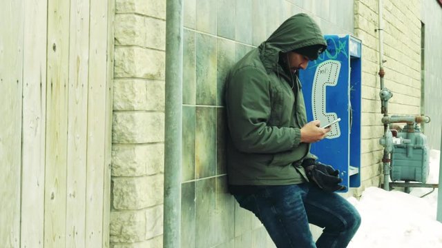 A poor man stands next to phonebooth playing on his cell phone