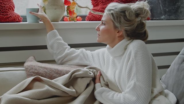 Young caucasian woman getting comfortable being carefree and smiling with warm tea or coffee on a couch with a cozy blanket during winter cold weather