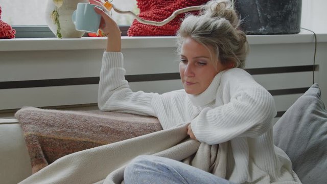 Cold almost ill woman getting comfortable underneath a cosy blanket with a cup of hot tea or coffee while trying to feel better