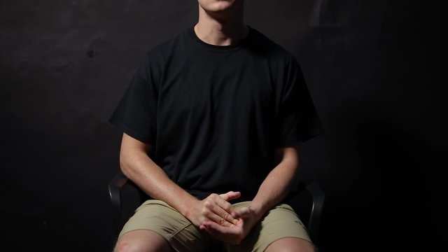 Slow motion shot of a man sitting on a chair fidgeting with his hands in anxiety. Shot in a studio with a black background.