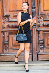 Obraz na płótnie Canvas Young South American Businesswoman working in New York City, wearing black sleeveless dress, arm carrying leather bag, walking dow stairs by brown vintage wooden office door, texting on cell phone..