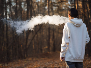 A man in a white hoodie exhales a cloud of steam smoke while walking in an autumn Park