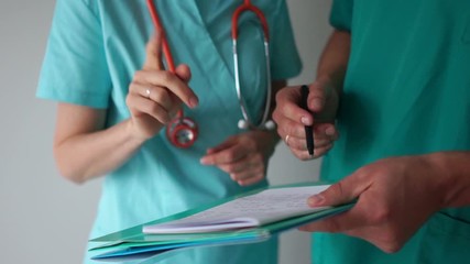 Man and woman in medical gown with stethoscope, folder and pen talking. Close-up of doctors hand discussing patient treatment plan