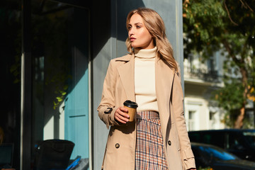 Beautiful stylish blond girl in beige coat with coffee to go walking on street