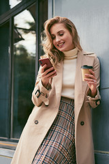 Beautiful smiling blond girl in trench coat with coffee to go happily talking on cellphone outdoor