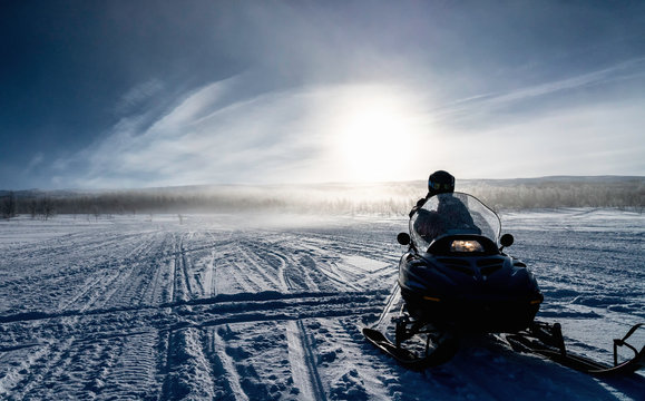 One person ready to drive snowmobile in very cold mountains in Sweden, frosty fog around bright sun creates halo effect, birches forest and mountains behind driver. Hemavan -Tarnaby area, Lappland