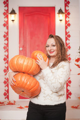 adult cute fat girl in a white sweater with gold stars holds a stack of large pumpkins and stands on the porch of her white house.