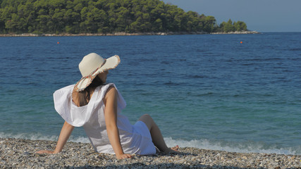 Fototapeta na wymiar Single woman with hat sit on beach sand and admire blue waiving sea, serenity
