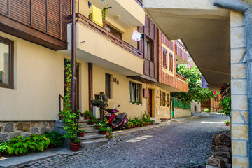 European narrow cobble-paved street between yellow houses in the old town. Small passage between brick buildings with flowers in pots near walls. Linen dries on rope. Red scooter parked near the door