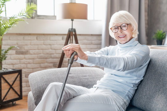 Smiling mature woman in glasses holding cane while sitting on sofa at home