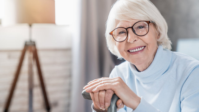 Portrait of happy mature woman in eyeglasses holding cane while sitting on sofa at home