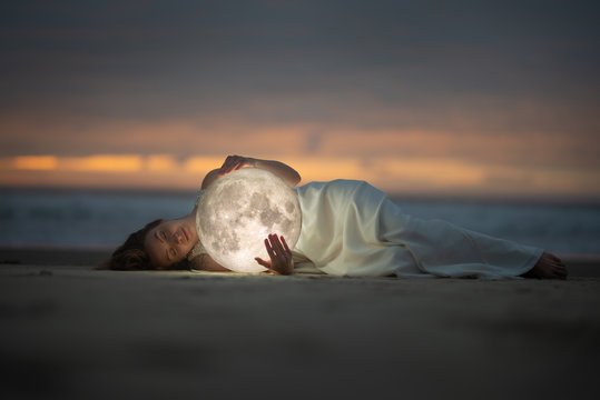 Astrology. Secret and riddle. Beautiful attractive girl on a night beach with sand hugs the moon, art photo.
