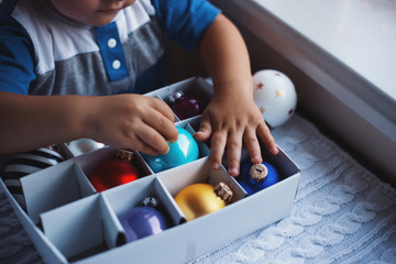 Happy child boy taking Christmas ball from box near window indoor with warm garland lights on blurred background.