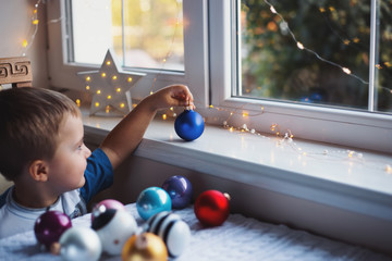 Happy little child boy holding blue Christmas ball near window indoor with warm garland lights on blurred background. - 296432223