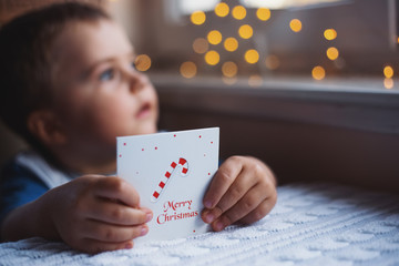 Child boy holding Christmas greeting card with warm garland bokeh on background near window in daylight. - 296432461