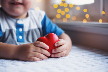 Smiling little child boy holding red toy heart in hands near window in daylight with cozy bokeh on background. The concept of love for children and cardiological care. - 296432636