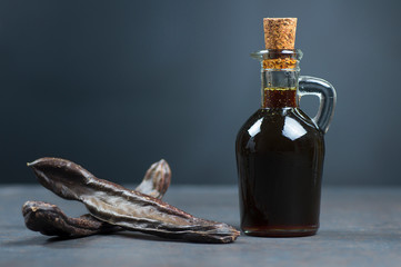 glass bottle of carob molasses and carob pods on rustic background, locust bean healthy food,...