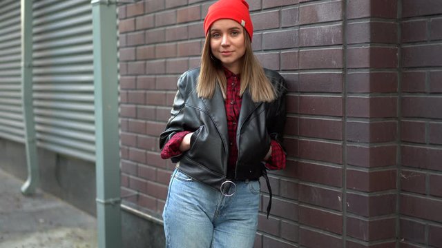 Beautiful European girl dressed black leather jacket and jeans and red hat standing backyard Hands in pockets leaning against red brick wall and smiling youth culture modern woman 30s year autumn 