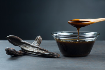 carob molasses in glass bowl and in wooden spoon and carob pods on rustic background, locust bean...