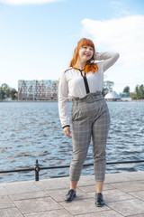 Cute happy young woman with red hair and business style clothes is standing on the shore of a city lake on a sunny day