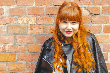 Cute young woman in a black leather jacket with curly red hair and bangs smiles against a brick...