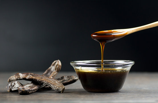 carob molasses in glass bowl and in wooden spoon and carob pods on rustic background, locust bean healthy food, Ceratonia siliqua ( harnup )