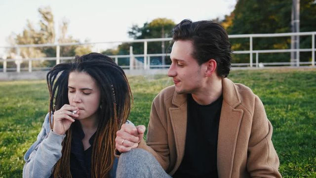 Happy young hipster couple sitting on grass and smoking marijuana joint in park, slow motion