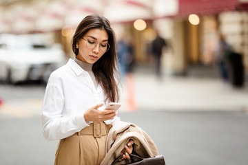 Beautiful business woman of Asian appearance standing on the street, typing a text message on her smartphone after a working day