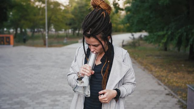 Portrait of young pretty woman with dreadlocks smoking marijuana with a bong in park